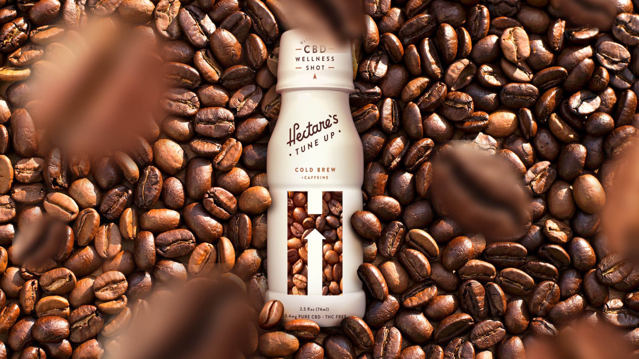 A bottle of Hēlteras Tune Up CBD Wellness Shot, Cold Brew flavor, is surrounded by coffee beans. The white bottle features minimalist branding and a window showing beans inside.