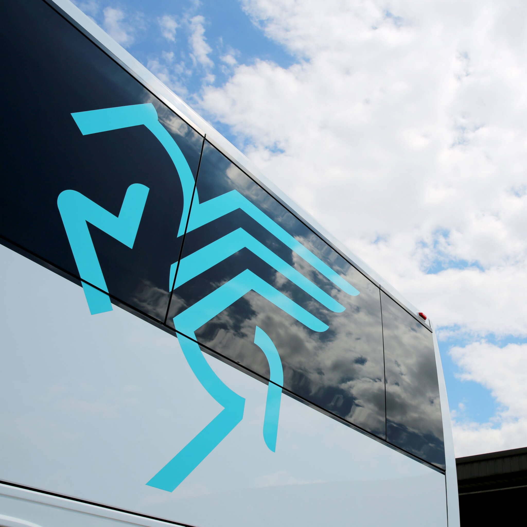 A modern bus with blue geometric graphics on its side is parked outside under a cloudy sky, reflecting clouds in its windows.