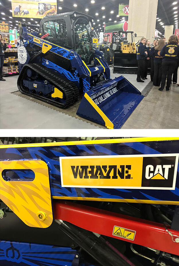 A blue and yellow CAT skid steer loader with “UNLOCK LIMITLESS” on the bucket, displayed indoors at an event. Close-up shows “WAYNE” and CAT logos on the side. People stand in the background.