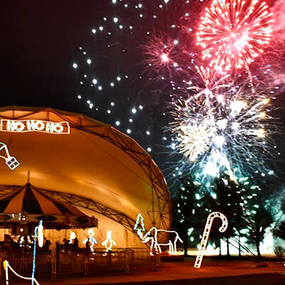 A festive night scene with colorful fireworks lighting up the sky above a dome-shaped stage. Holiday lights shaped like a candy cane, horse, and HO HO HO decorate the area, creating a cheerful atmosphere.