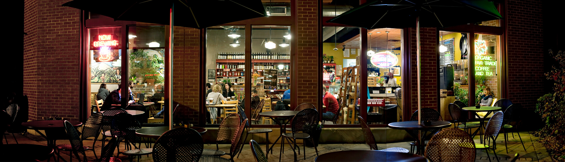 A nighttime view of a cozy café with large windows, showing people sitting at tables inside. Outdoor tables and chairs are empty, illuminated by streetlights and neon signs, with shelves and decor visible through the windows.