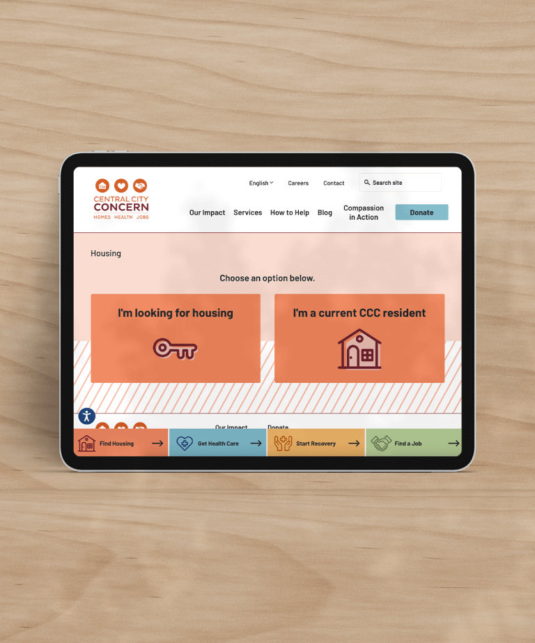 Tablet displaying Central City Concern website with options: Im looking for housing (key icon) and Im a current CCC resident (house icon), against a light wood background.