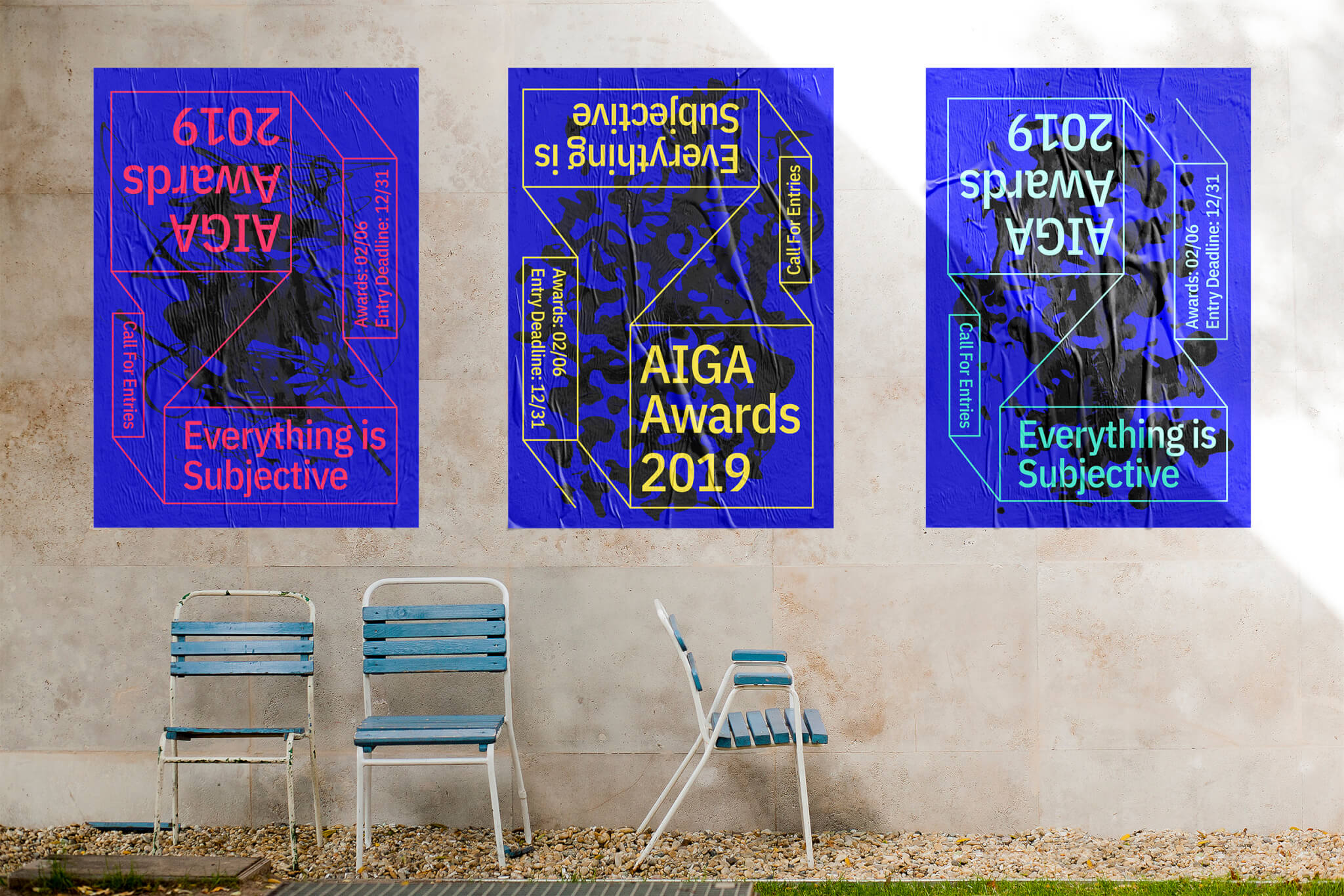 Three colorful posters for the AIGA Awards 2019 with the phrase “Everything is Subjective” are displayed on a wall above three empty blue and white chairs on a gravel surface.
