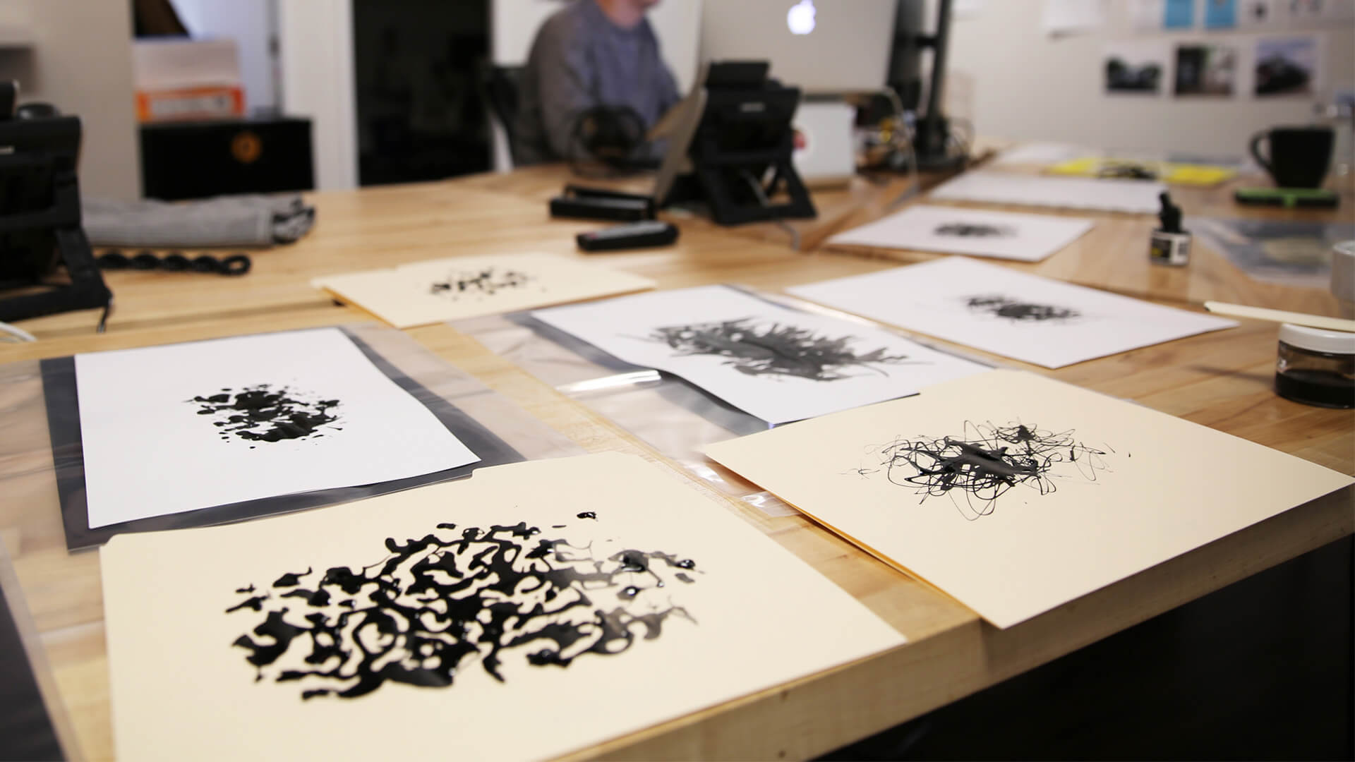 Several abstract black ink drawings are spread out on a wooden table in a creative studio, with art supplies and a blurred figure working at a desk in the background.