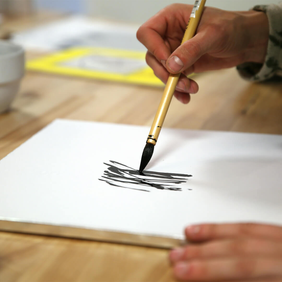 A person holding a paintbrush draws black strokes on white paper, with a wooden table and a cup in the background.
