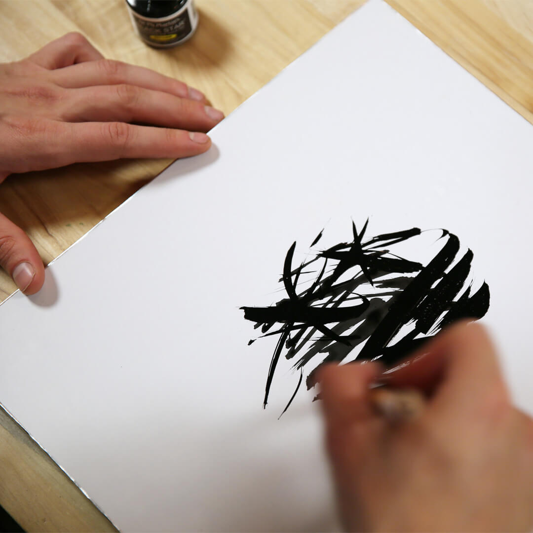 A person is using a brush and black ink to create abstract, bold strokes on a white sheet of paper placed on a wooden surface.