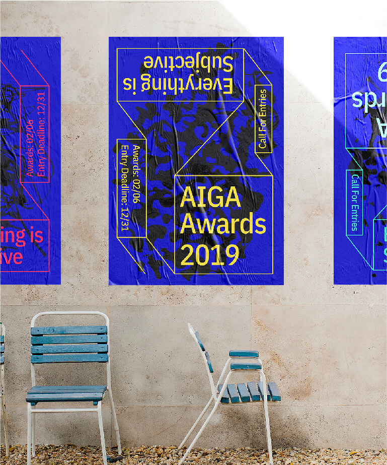 A blue AIGA Awards 2019 poster with yellow and white text on a wall next to two blue and white metal chairs. The poster has geometric lines and a tiger graphic in the background.
