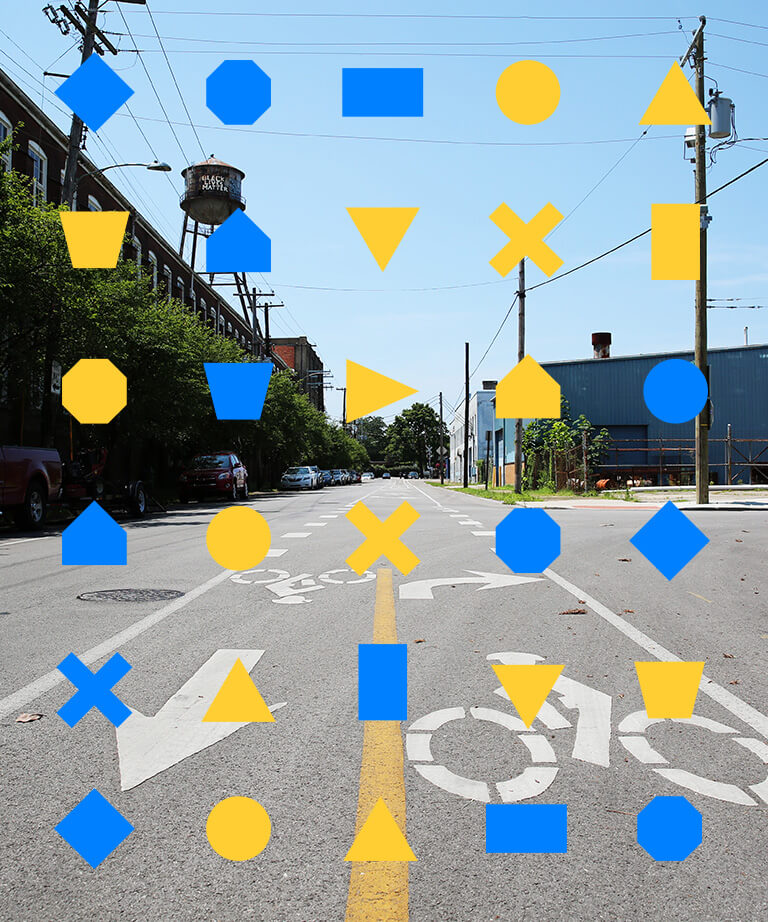 A city street with bike lanes is overlaid by a grid of blue and yellow geometric shapes, including circles, triangles, squares, and octagons. Trees, buildings, and a water tower are visible in the background.