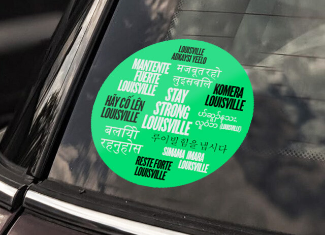 A round green car sticker on a window reads Stay Strong Louisville in multiple languages, surrounded by translations and phrases of encouragement in different scripts and fonts.