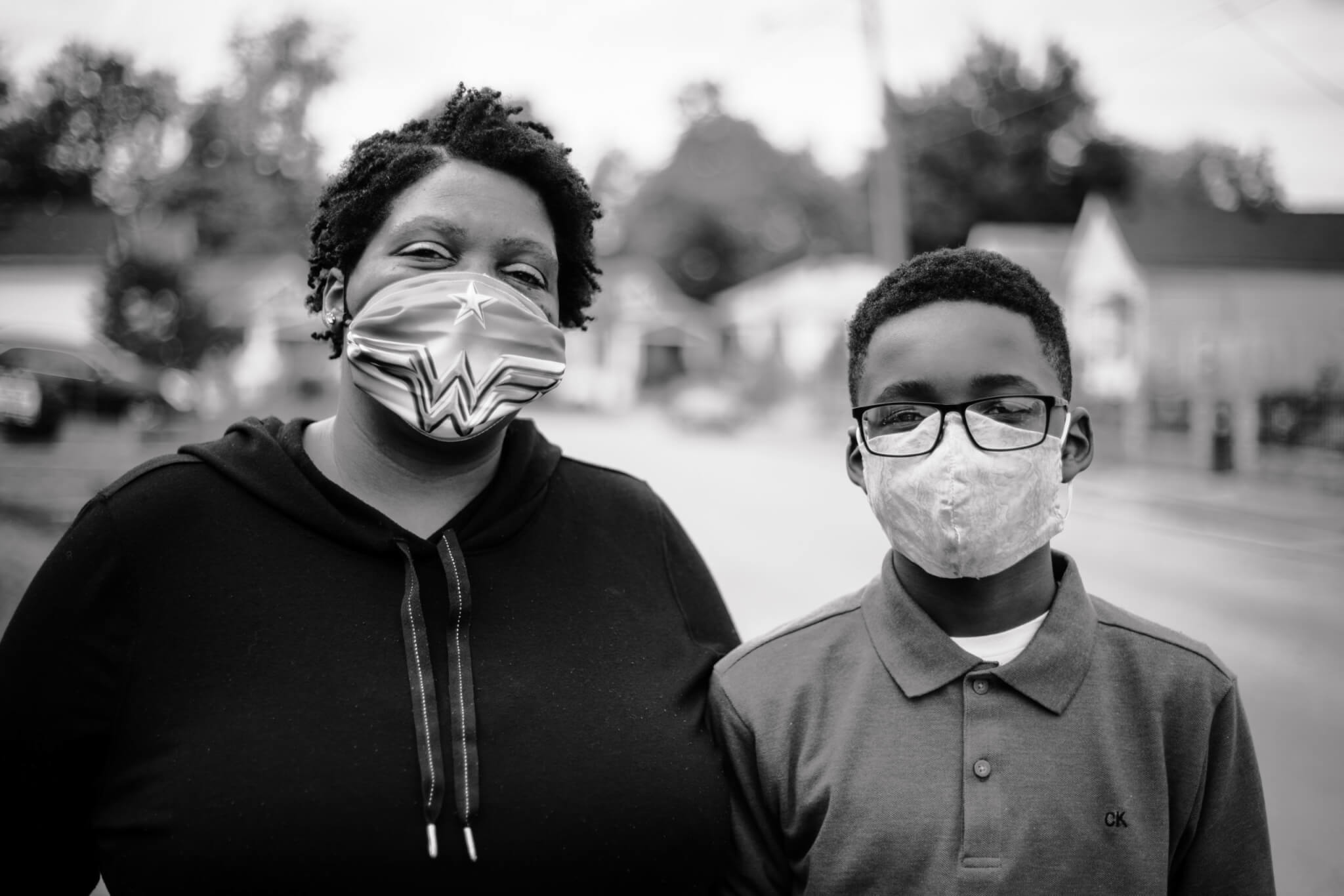A woman and a boy wearing face masks stand side by side outdoors on a street, looking at the camera. The woman’s mask has a Wonder Woman design. Both appear calm and the background is slightly blurred.