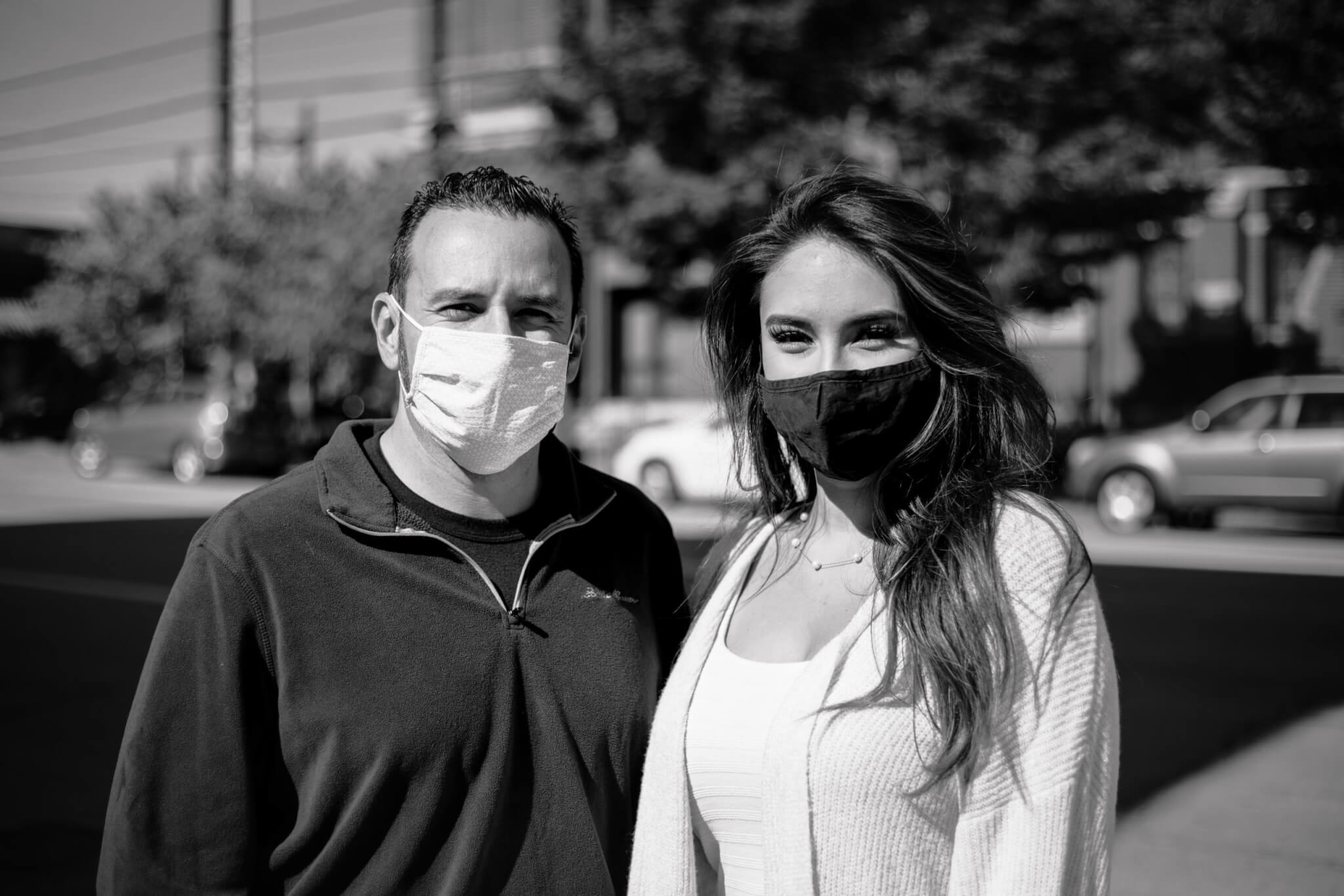 A man and a woman wearing face masks stand close together outdoors on a sunny day, with cars and trees visible in the blurred background. The image is in black and white.