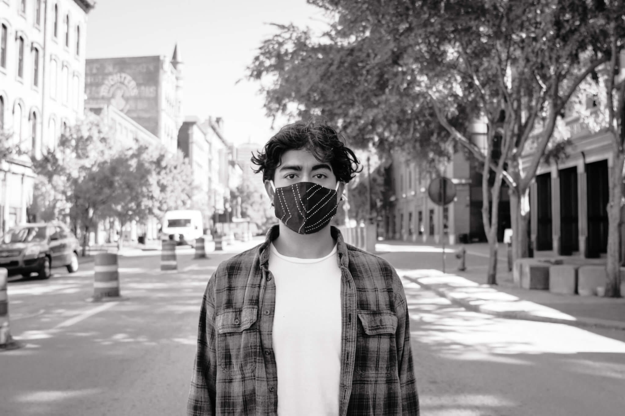 A young person wearing a patterned face mask and plaid shirt stands in the middle of an empty city street lined with buildings and trees. The image is in black and white.