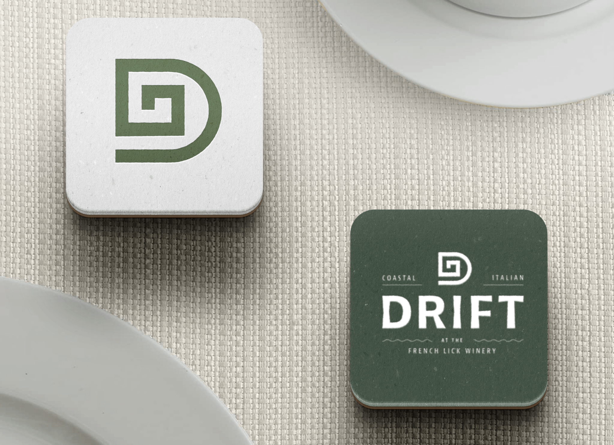 Two square coasters on a textured beige surface; one features a stylized green D logo, the other displays the same logo above the text DRIFT at the French Lick Winery with white dishes nearby.