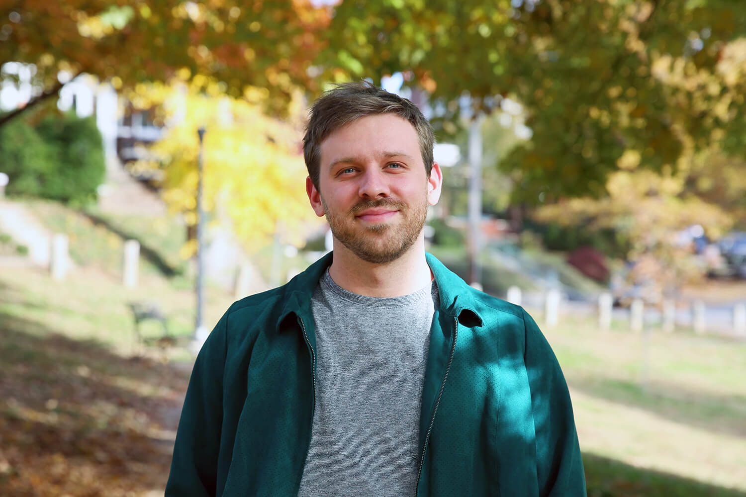 A man with short hair and a beard stands outdoors under autumn trees, wearing a gray shirt and green jacket, smiling softly at the camera. The background is filled with colorful fall foliage.