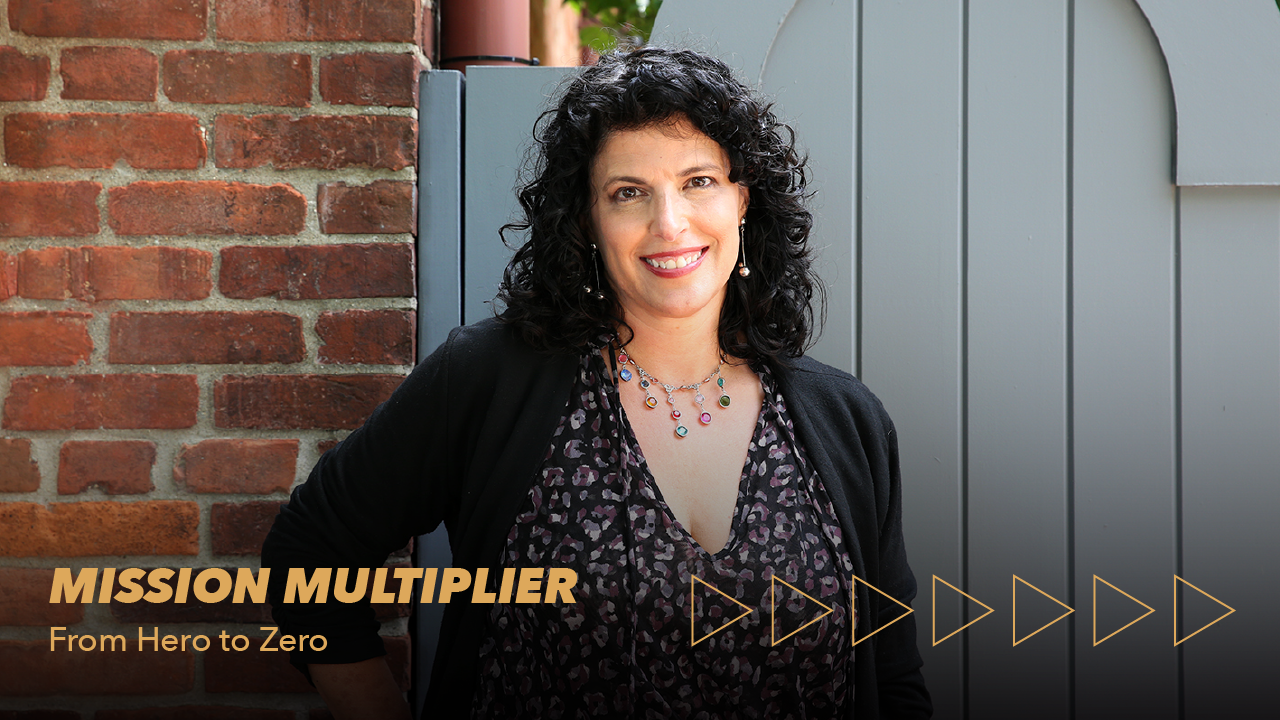 A smiling woman with curly dark hair stands by a brick wall and gray gate. Text reads MISSION MULTIPLIER From Hero to Zero with gold arrow graphics on the lower right.