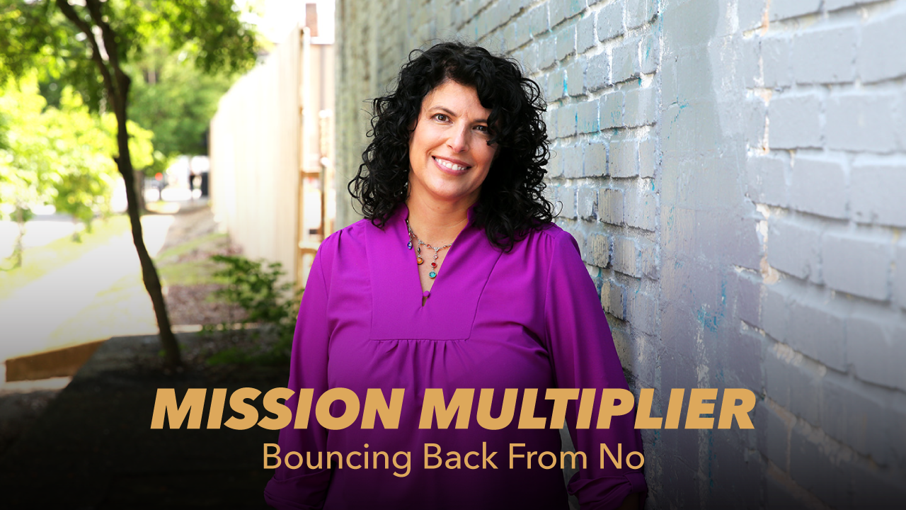 A woman with curly dark hair, wearing a bright purple blouse, stands smiling beside a light blue brick wall outdoors. Text reads: MISSION MULTIPLIER—Bouncing Back From No.