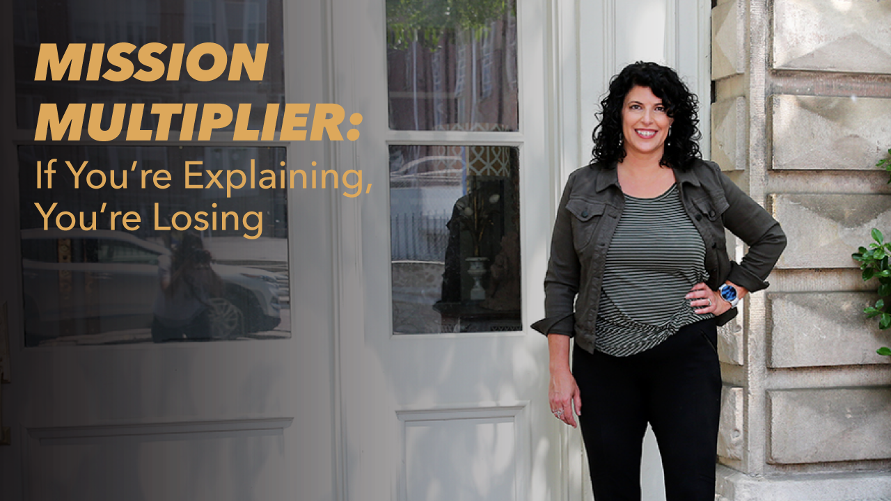 A woman with curly dark hair stands smiling by a building entrance. Text on the image reads: MISSION MULTIPLIER: If You’re Explaining, You’re Losing.