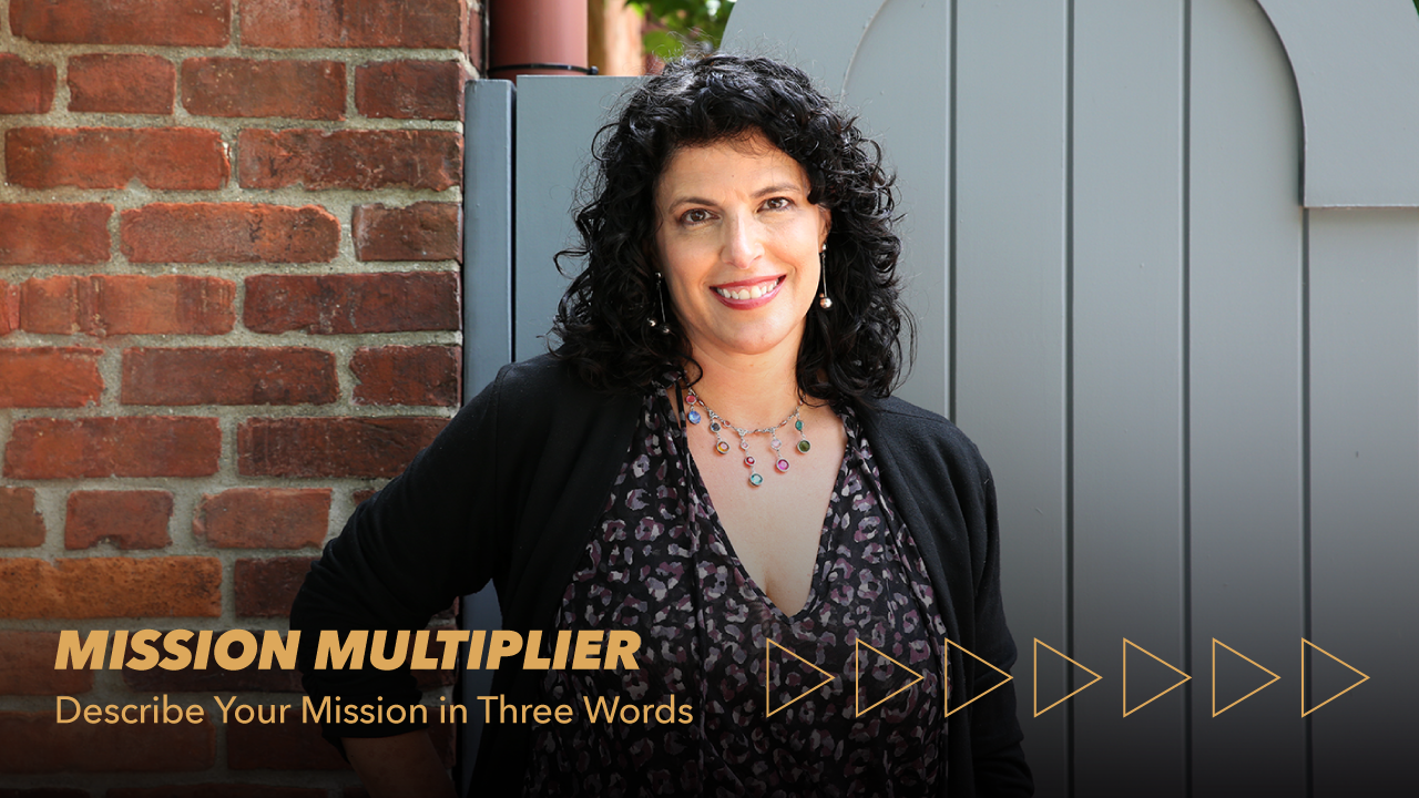 A woman with curly dark hair stands smiling near a brick wall and gray gate. Text reads: MISSION MULTIPLIER - Describe Your Mission in Three Words with arrow graphics below.