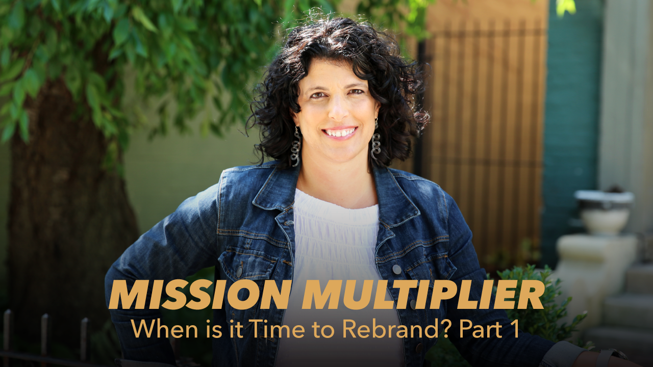 A woman with curly dark hair smiles outdoors, wearing a denim jacket. Overlaid text reads: “MISSION MULTIPLIER When is it Time to Rebrand? Part 1.” Green foliage and a building are in the background.