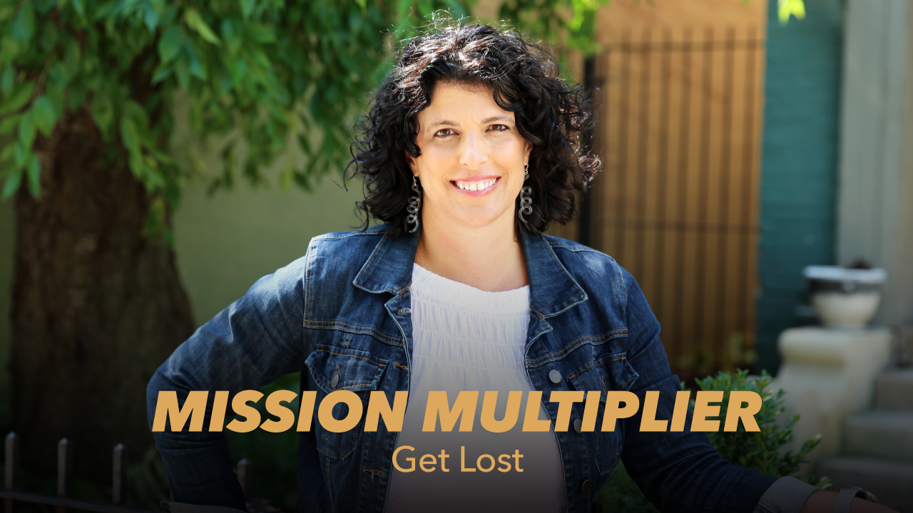 A smiling woman with curly dark hair stands outdoors wearing a denim jacket. The text MISSION MULTIPLIER Get Lost is overlaid at the bottom of the image. Greenery and a building are in the background.