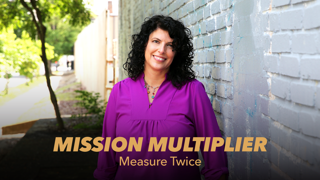 A woman with curly dark hair, wearing a purple blouse, stands smiling beside a sunlit brick wall. Text on the image reads: MISSION MULTIPLIER Measure Twice.