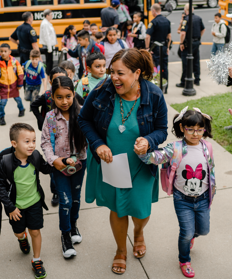 A smiling woman holding a paper leads a group of young children, some holding hands, as they walk together on a sidewalk near a school bus. Other children and adults are visible in the background.
