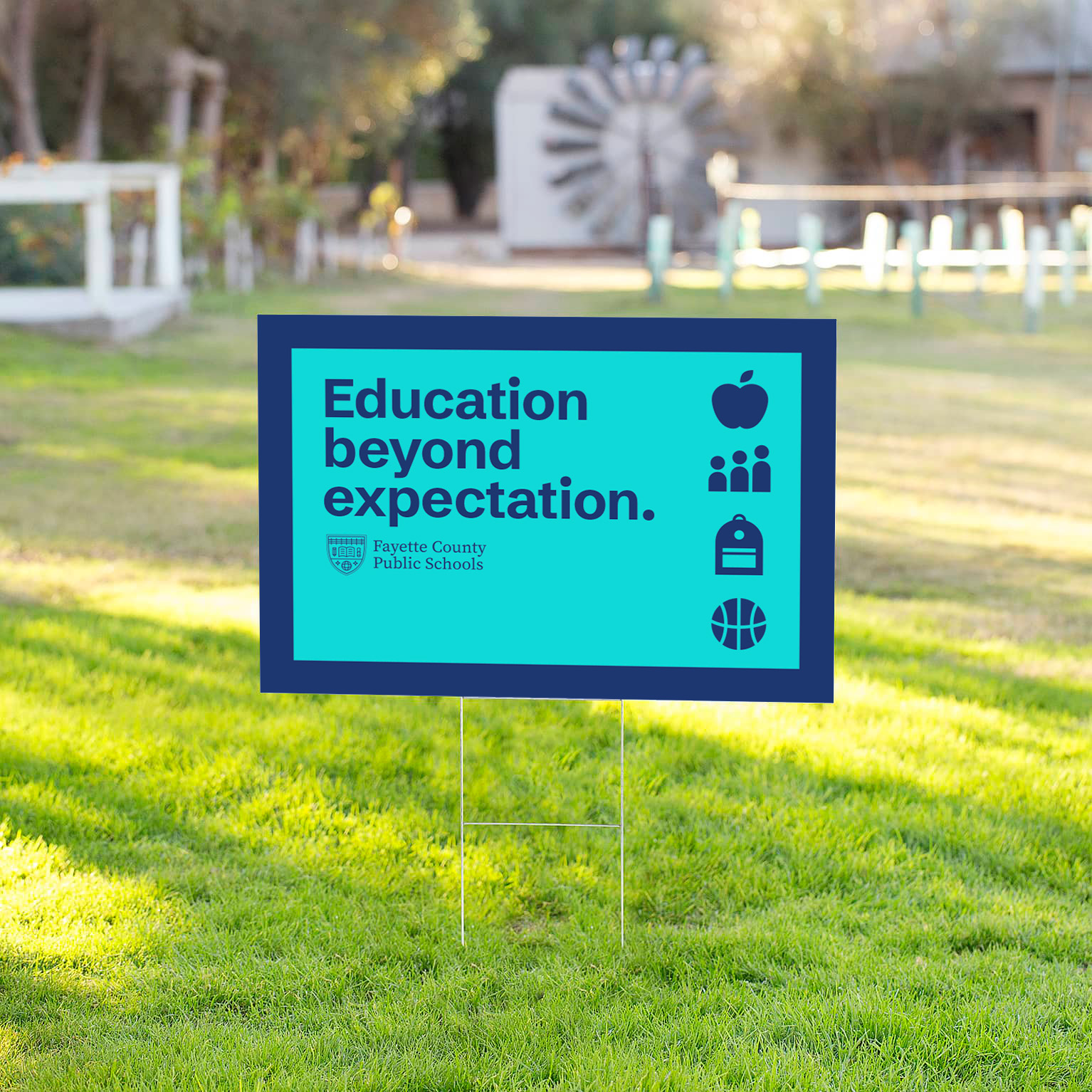 A blue Fayette County Public Schools yard sign reads Education beyond expectation with icons of an apple, a lock, a basketball, and people, displayed on green grass in a sunny outdoor area.