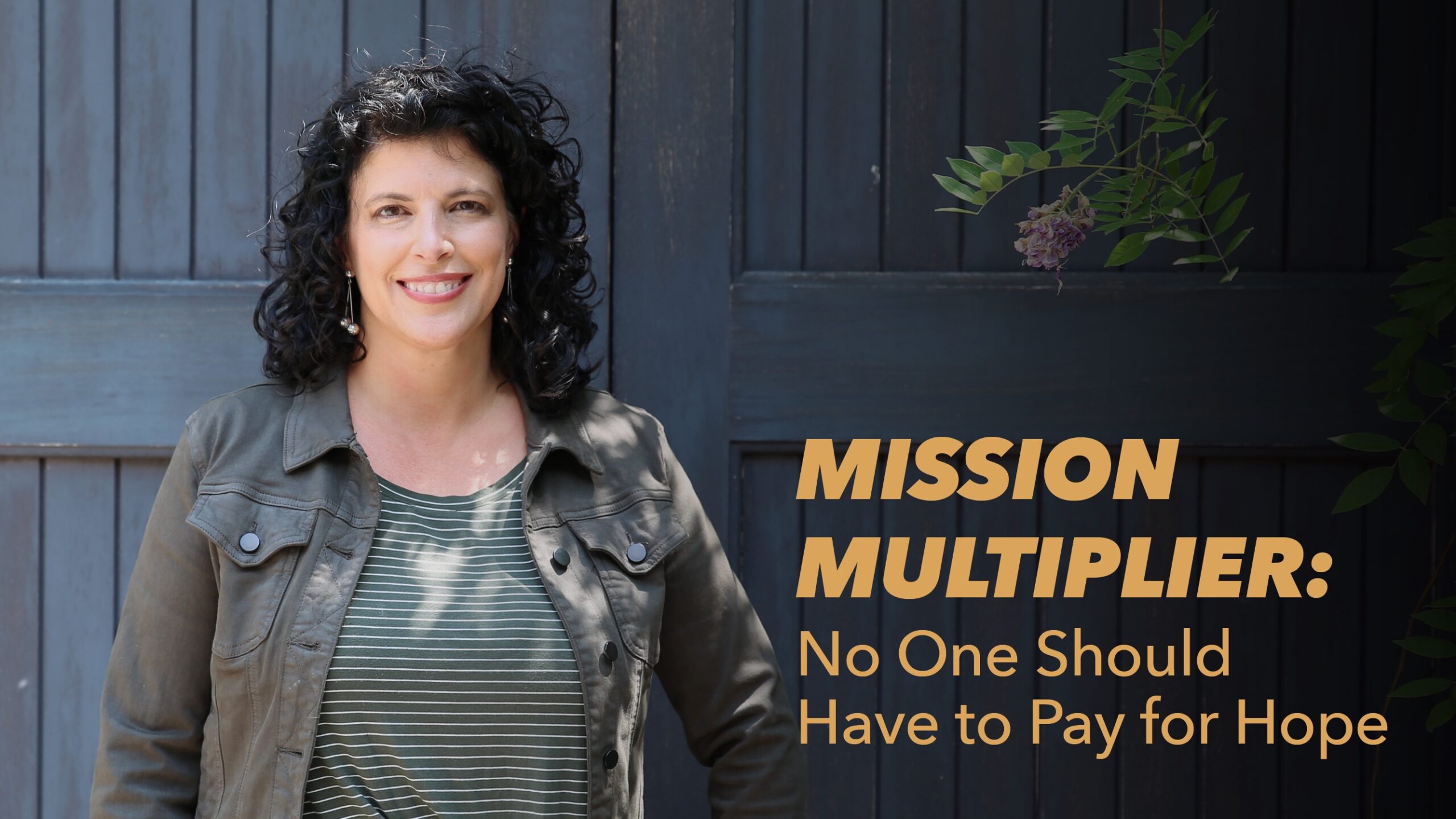 A woman with curly dark hair, wearing a striped shirt and olive jacket, stands smiling in front of a dark wooden wall. Text reads: Mission Multiplier: No One Should Have to Pay for Hope.