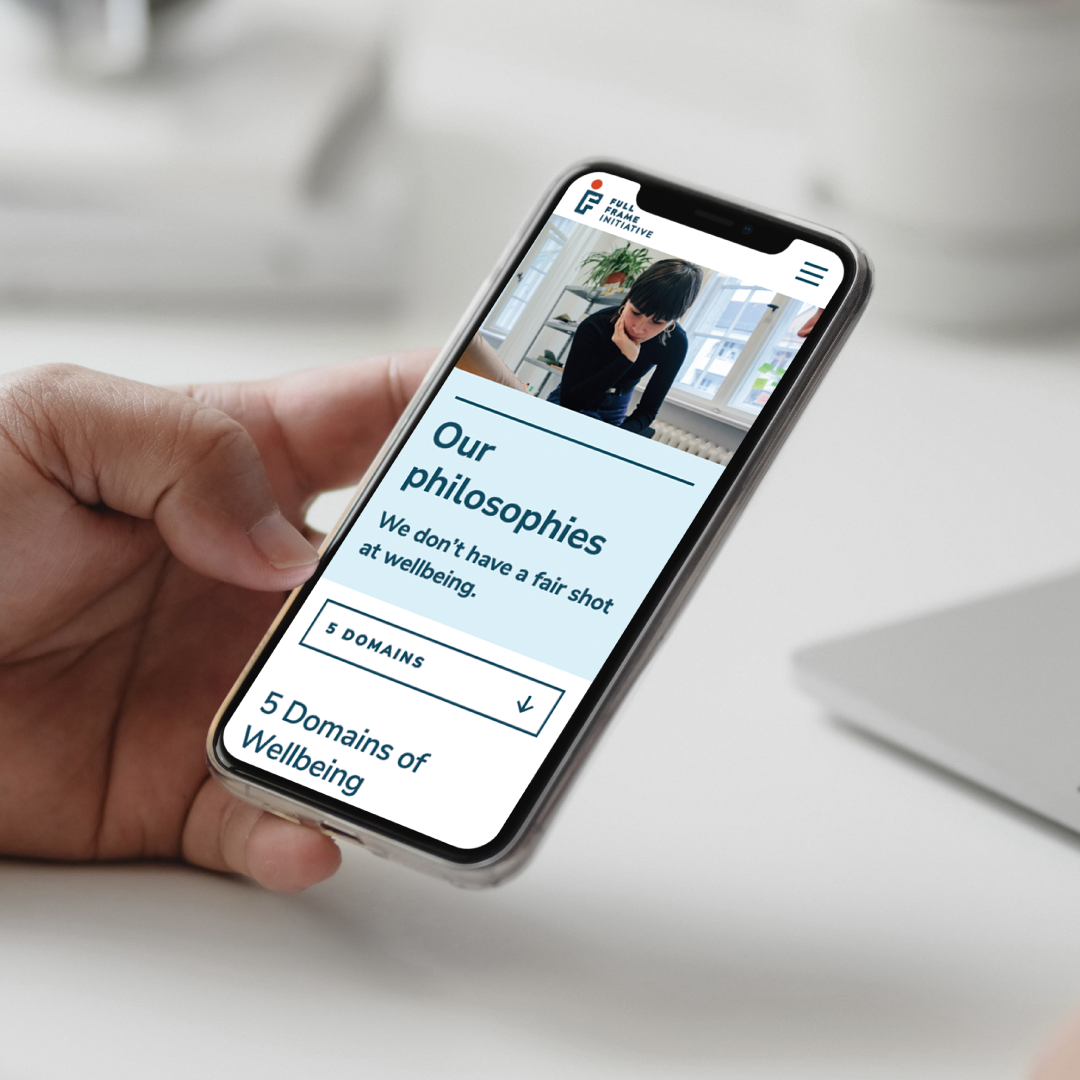 A hand holds a smartphone displaying a website page titled Our philosophies with the subtitle We dont have a fair shot at wellbeing and a photo of a person sitting and writing at a desk.