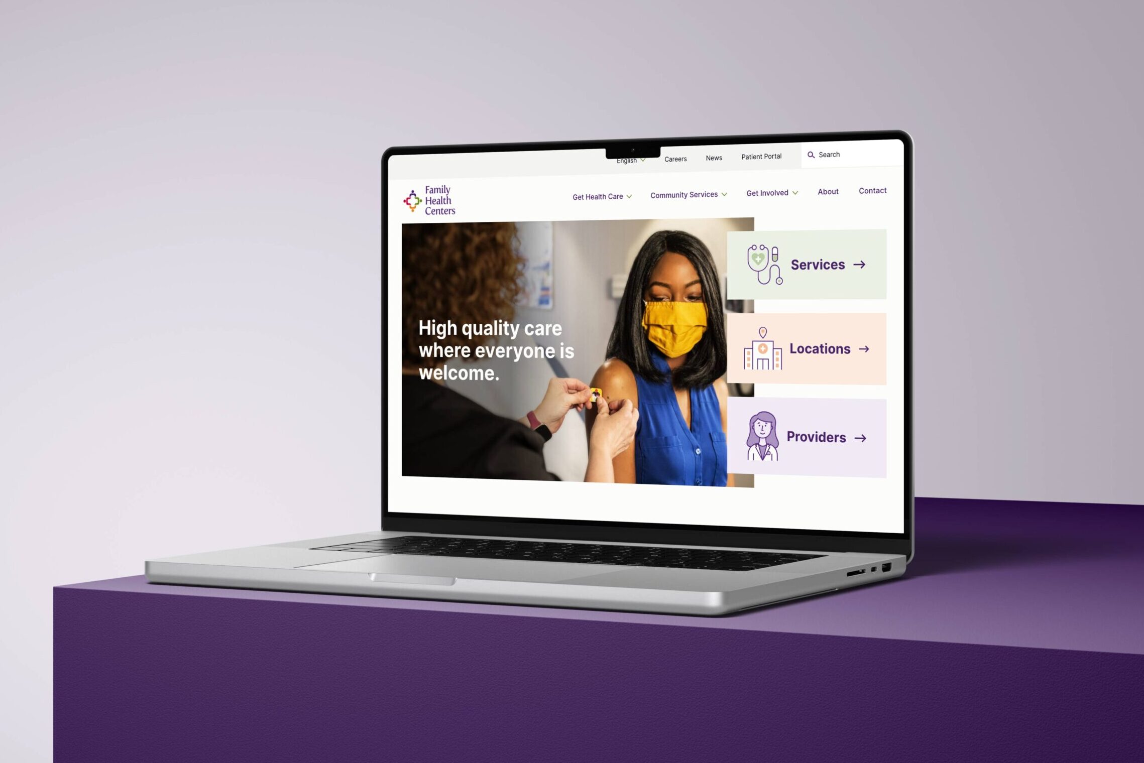 A laptop displays a health center website. The screen shows a woman in a yellow mask receiving a vaccine. Text reads High quality care where everyone is welcome with menu options for Services, Locations, and Providers.