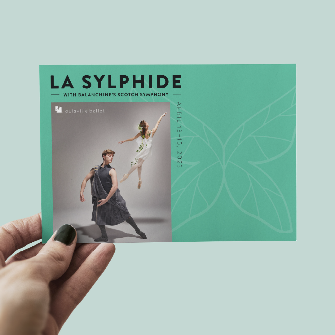 A hand holds a teal postcard for La Sylphide with Balanchine’s Scotch Symphony, featuring two ballet dancers and the Louisville Ballet logo. Event dates, April 13–15, 2023, are listed along the side.