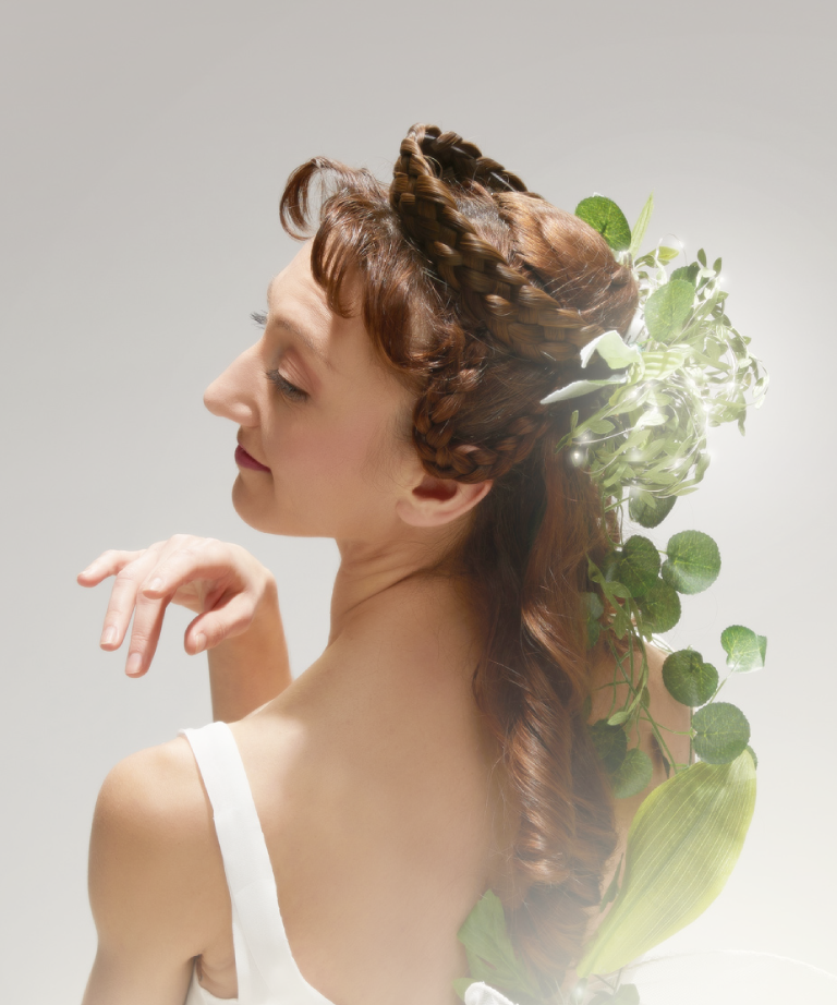 A woman in a white dress poses gracefully with her head tilted and eyes closed. Her long brown hair is styled with braids and adorned with green leaves and plants. The background is light and neutral.