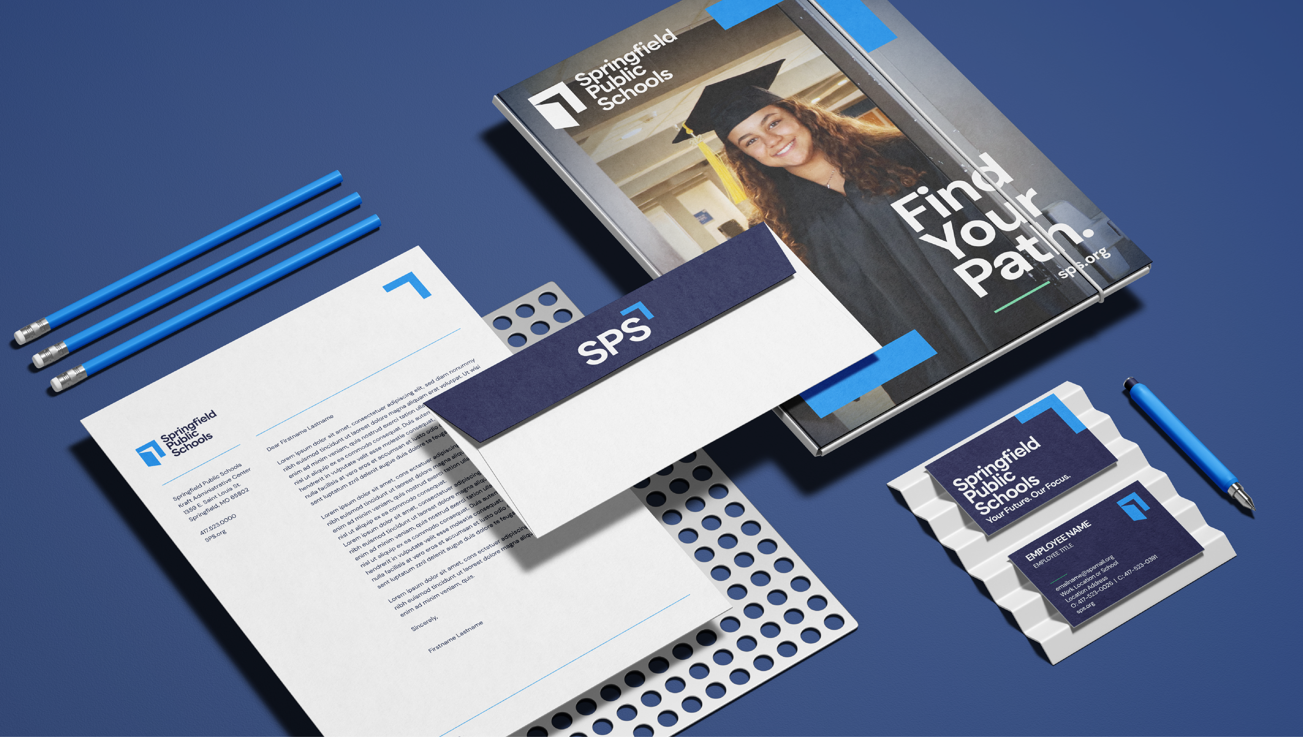 A flat lay of branded stationery for Springfield Public Schools on a blue background, including a letter, envelope, brochures, business cards, pencils, a pen, and a notebook with a students graduation photo and the text “Find Your Path.”.