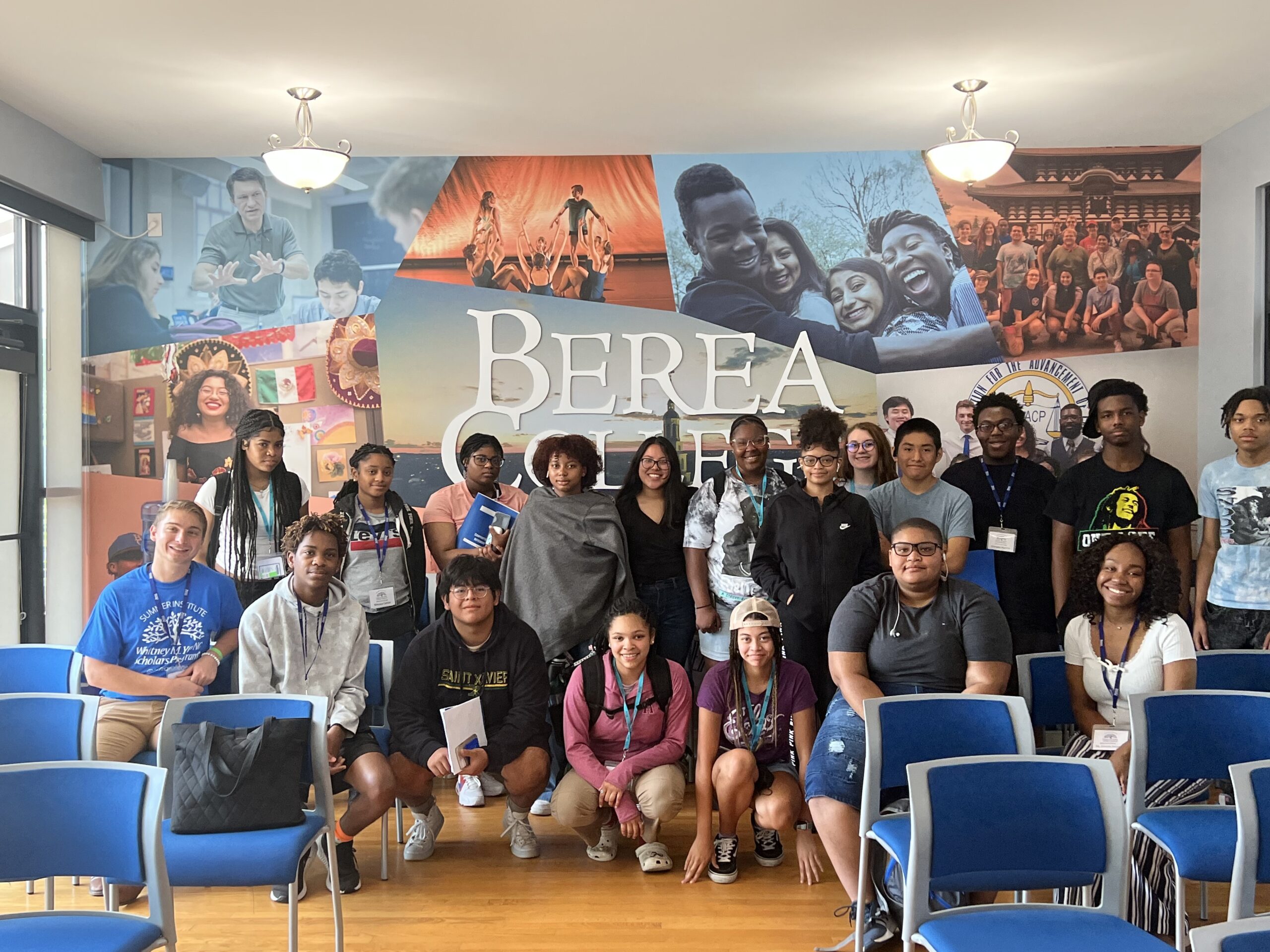 A diverse group of around 20 high school or college-age students pose for a group photo in a bright room at Berea College. They are smiling and standing or sitting in front of a large wall mural that features the Berea College logo and images of student life, including classroom scenes, group hugs, and cultural events. Blue chairs are arranged in rows in the foreground.